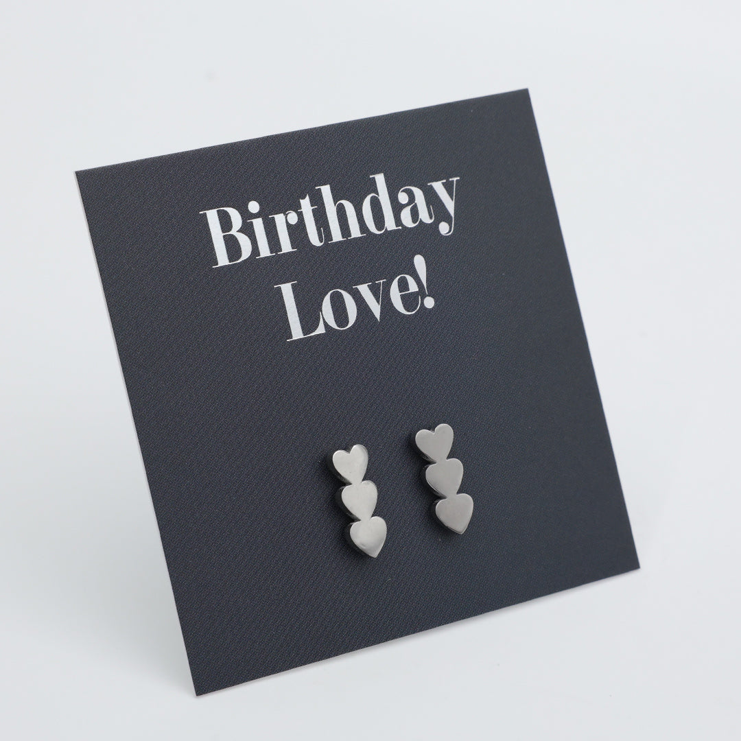 Three pairs of small silver heart stud earrings displayed on a dark charcoal square card that reads 'Birthday Love!' in white text, photographed on a light gray background.