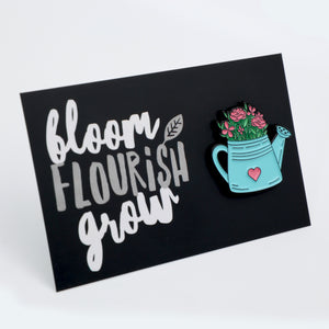 A rectangular black sign featuring the words 'bloom FLOURISH grow' in white and grey lettering, alongside a turquoise watering can with pink flowers and a small pink heart.