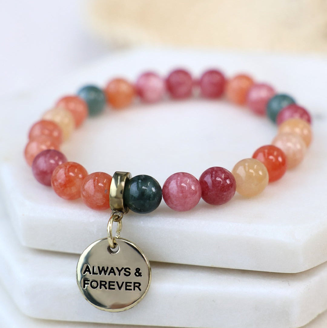 A beaded stretch bracelet with round gemstone-like beads in pink, orange, red, and dark green, featuring a gold-tone charm engraved with 'ALWAYS & FOREVER', displayed on stacked white stone blocks.