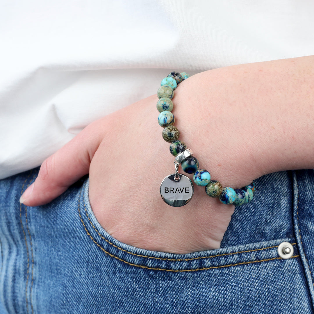 Bracelet with 'BRAVE' charm on wrist, wearing jeans and a white shirt