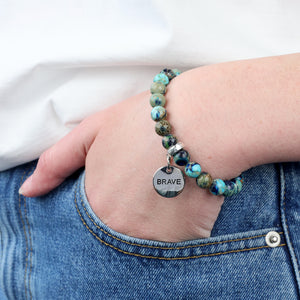 Bracelet with 'BRAVE' charm on wrist, wearing jeans and a white shirt