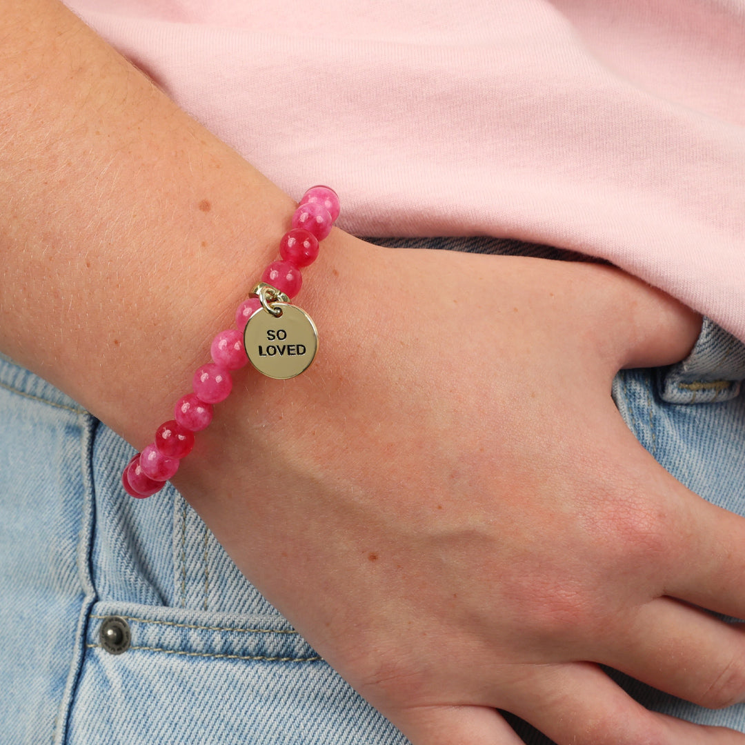 Pink beaded bracelet with a gold tag on a wrist, against a pink shirt and blue jeans background.
