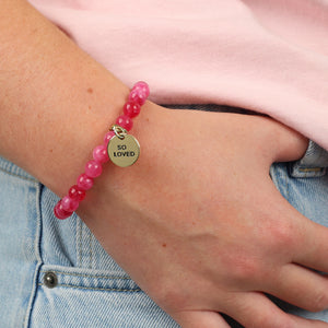 Pink beaded bracelet with a gold tag on a wrist, against a pink shirt and blue jeans background.