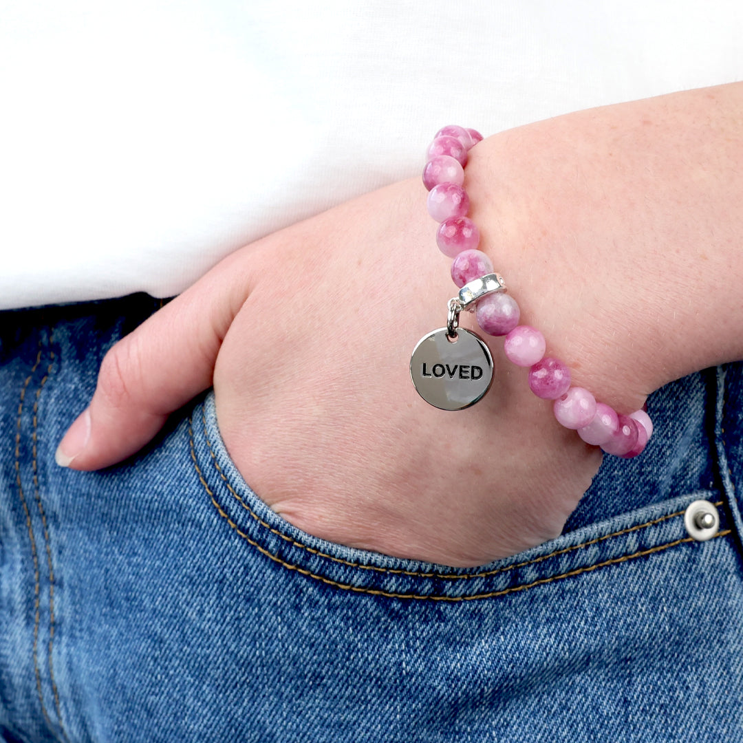 Pink beaded bracelet with a 'LOVED' charm on a wrist, worn with blue jeans.