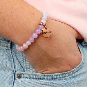 Bracelet with pink beads and a gold tag labeled 'CHERISH' on a wrist.