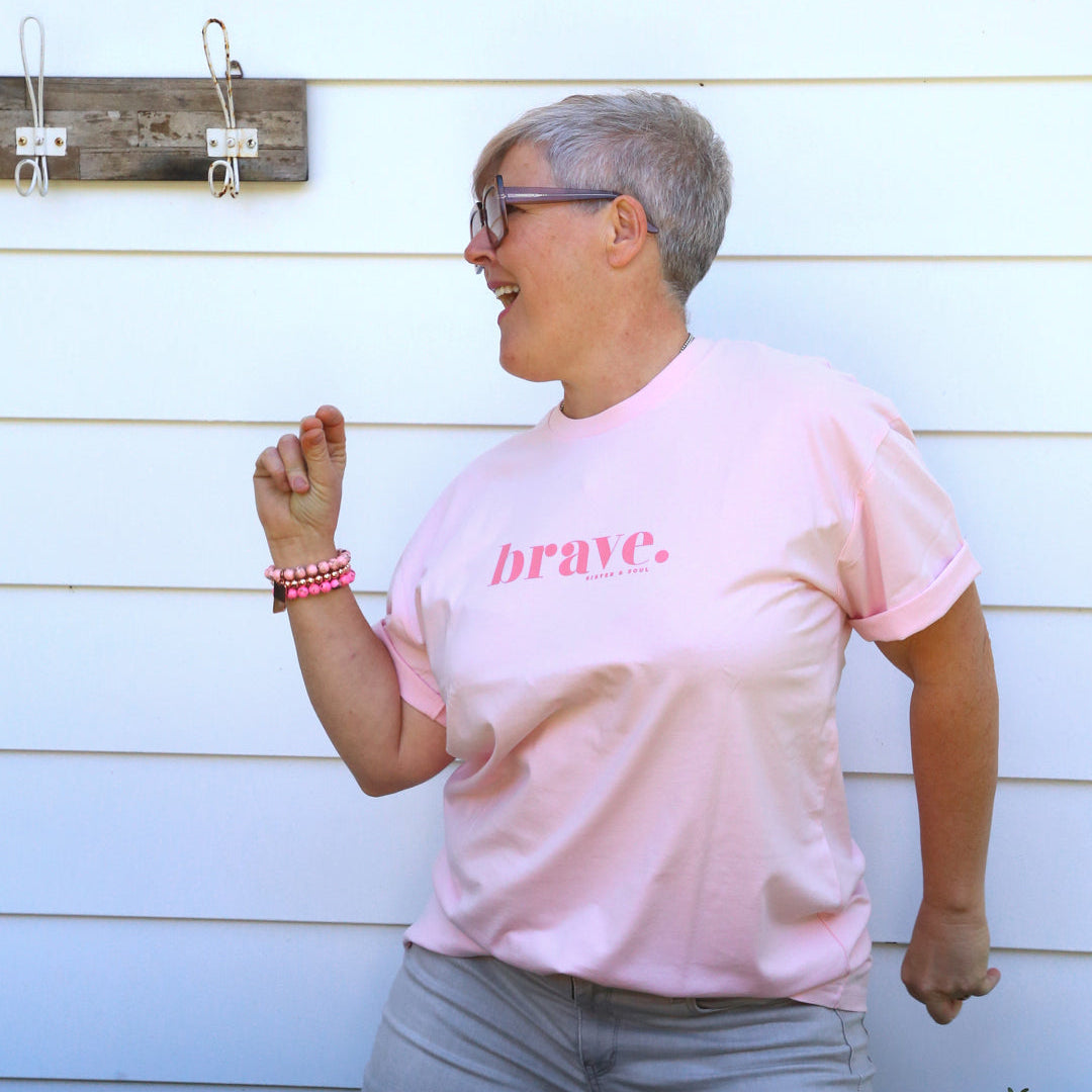 A woman with short gray hair and glasses wearing a light pink t-shirt with the word 'brave.' printed in dark pink on the chest, paired with gray pants, standing in front of a white house exterior.