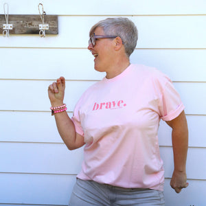 A woman with short gray hair and glasses wearing a light pink t-shirt with the word 'brave.' printed in dark pink on the chest, paired with gray pants, standing in front of a white house exterior.