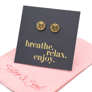 Gold shell earrings on a card with 'breathe. relax. enjoy.' text, placed on a pink fabric background.