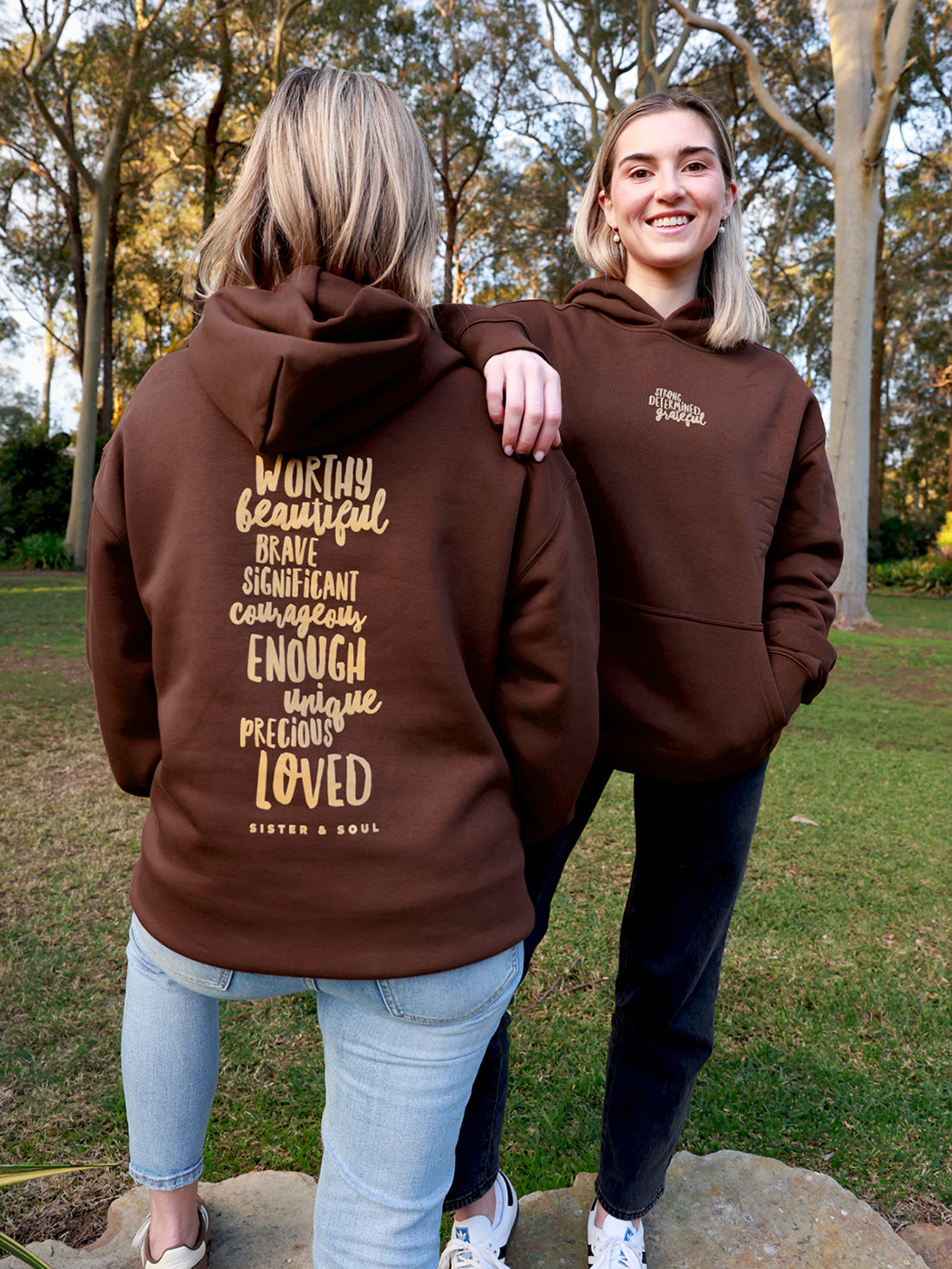 Two women outdoors in a park wearing brown hoodies. The woman on the left shows the back of the hoodie with a large gold typographic quote reading 'Worthy beautiful brave significant enough ENOUGH LOVED' and 'SIXTEN AND SOUL'.