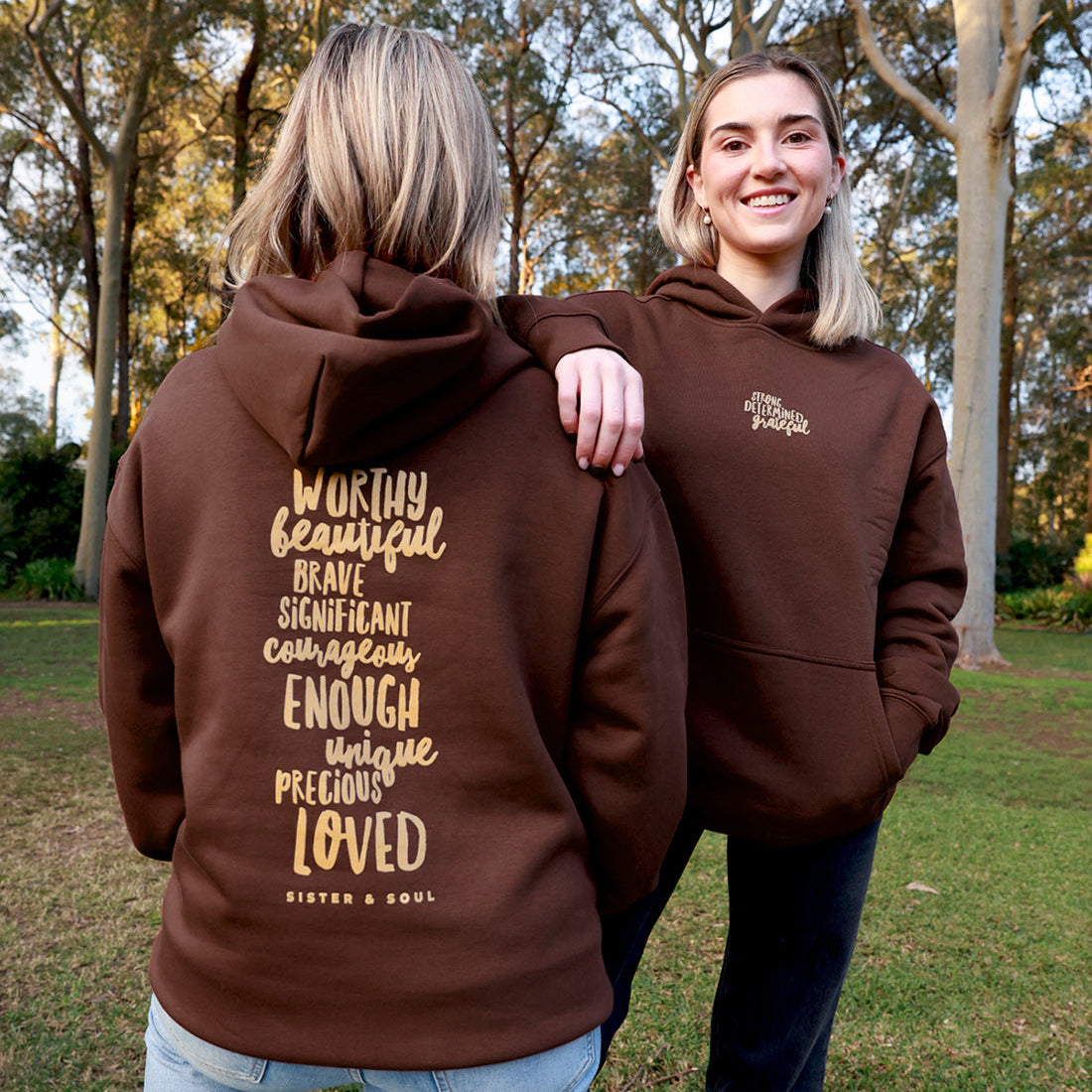 Two women outdoors in a park wearing brown hoodies. The woman on the left shows the back of the hoodie with a large gold typographic quote reading 'Worthy beautiful brave significant enough ENOUGH LOVED' and 'SIXTEN AND SOUL'.
