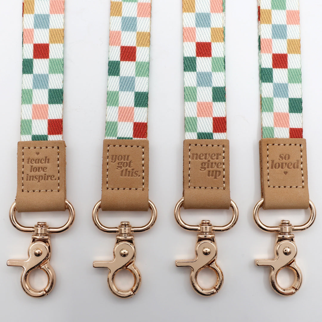 Four checkered lanyards with brown leather tags and rose gold clasps on a light gray background.