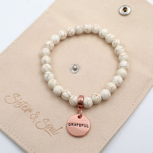 White beaded bracelet with a rose gold 'GRATEFUL' charm on a beige card with 'Sister & Soul' branding.