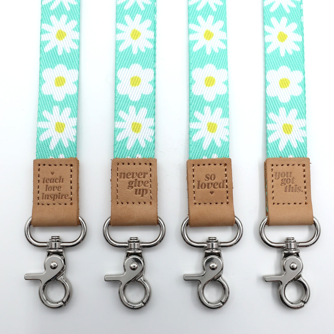 Four keychains with daisy pattern straps and motivational text on brown leather tabs against a white background.