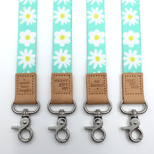 Four keychains with daisy pattern straps and motivational text on brown leather tabs against a white background.