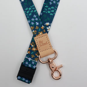 Be Kind embossed on a dark blue floral lanyard with tan trim and a lobster clasp.