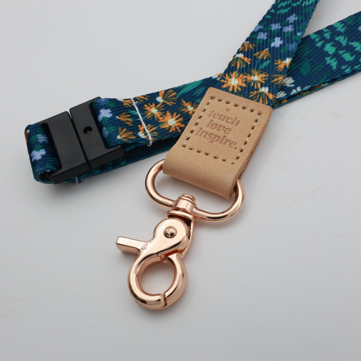 Teach Love Inspire embossed on a dark blue floral lanyard with tan trim and a lobster clasp.