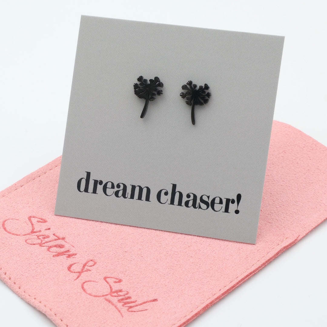 Black earrings on a card with 'dream chaser!' text, placed on a pink surface with 'Sister & Soul' branding.
