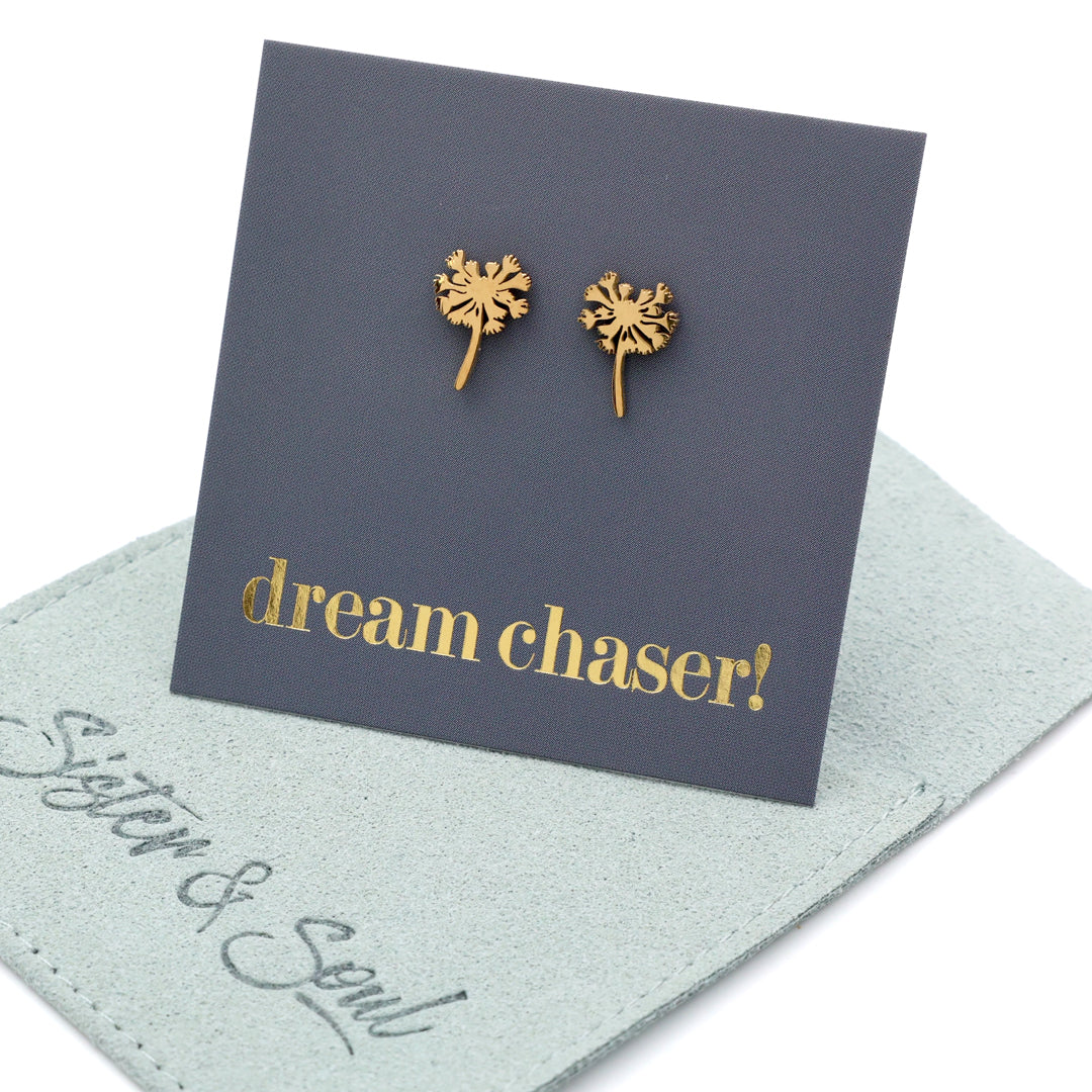 Gold dandelion-shaped earrings on a card with 'dream chaser!' text, displayed on a light blue mat.