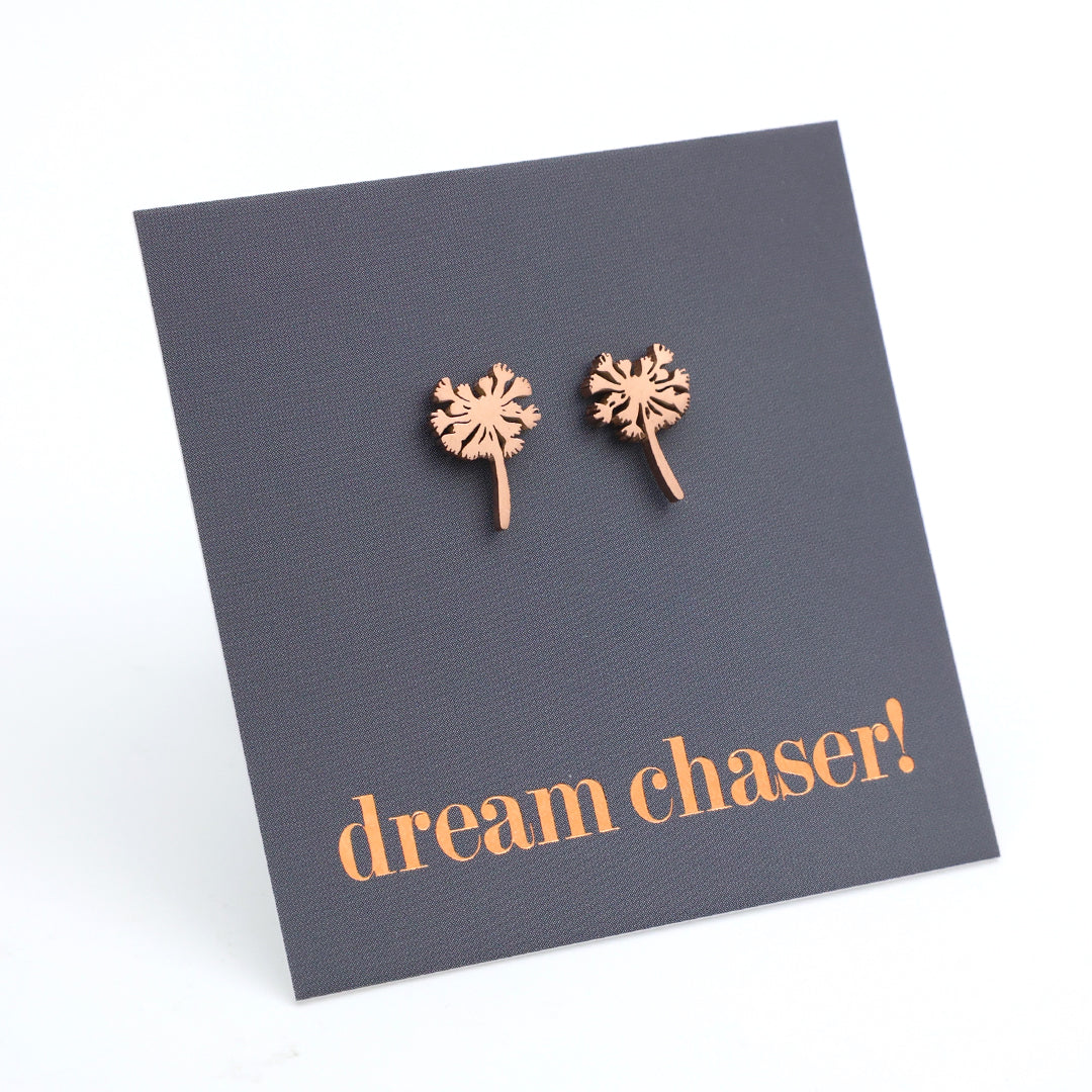 Rose gold dandelion-shaped earrings on a dark gray card with 'dream chaser!' text.