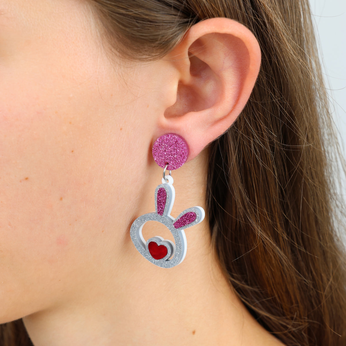 A pair of acrylic statement dangle earrings in the shape of bunny faces with pink glittery tops and red hearts, displayed on a black background with the text 'SO LOVED'.