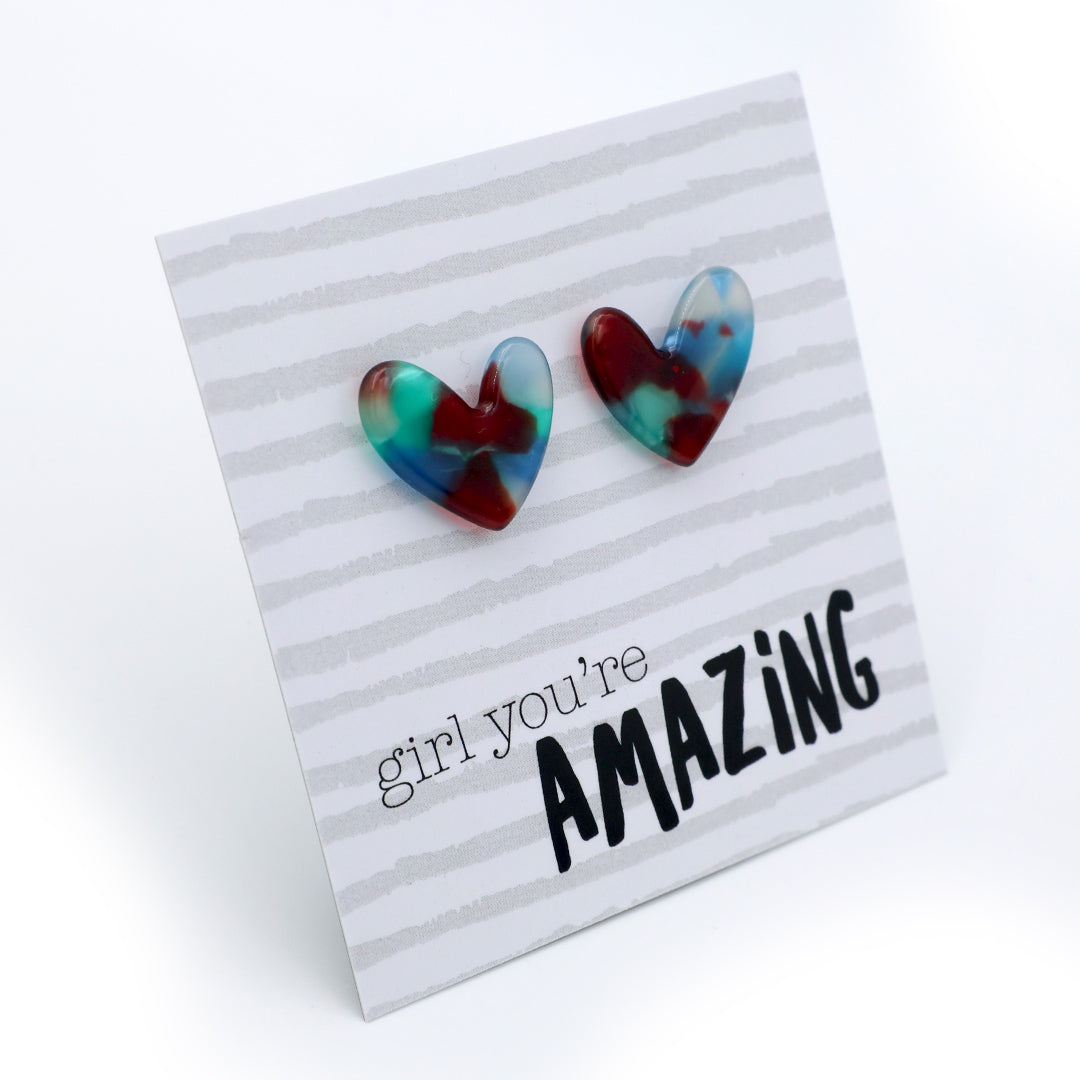 A pair of heart-shaped stud earrings with a marbled swirl pattern of red, blue, and green, displayed on a white card with the text 'girl you're AMAZING' printed on it.