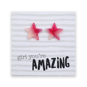 A pair of star-shaped stud earrings in a pink ombre gradient, displayed on a white card with light gray horizontal stripes. The card features the printed text 'girl you're AMAZING' in black and gray fonts.