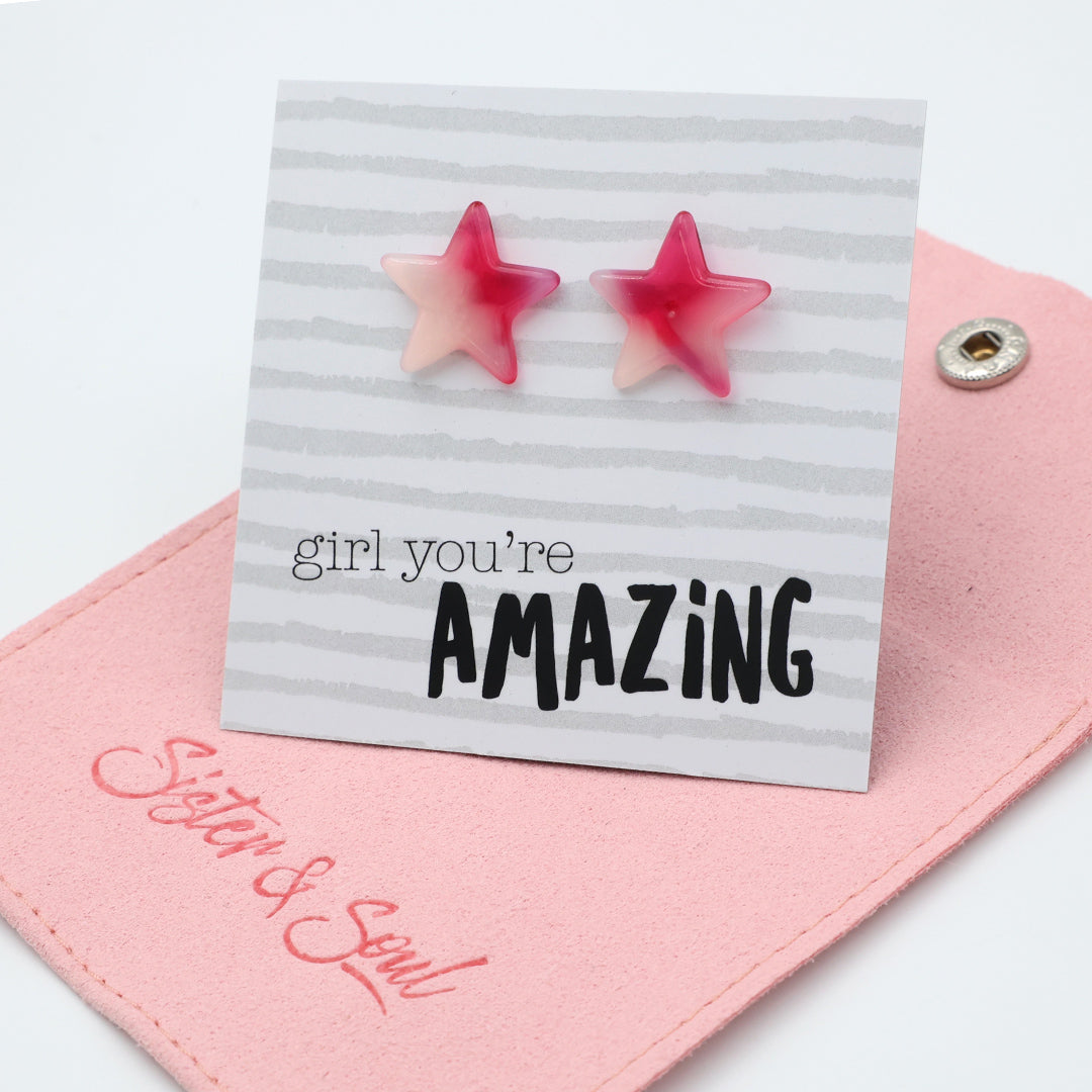 A pair of pink and red star-shaped stud earrings displayed on a white striped card that reads 'girl you're AMAZING', resting on a pink fabric pouch with ' Sister & Soul'.