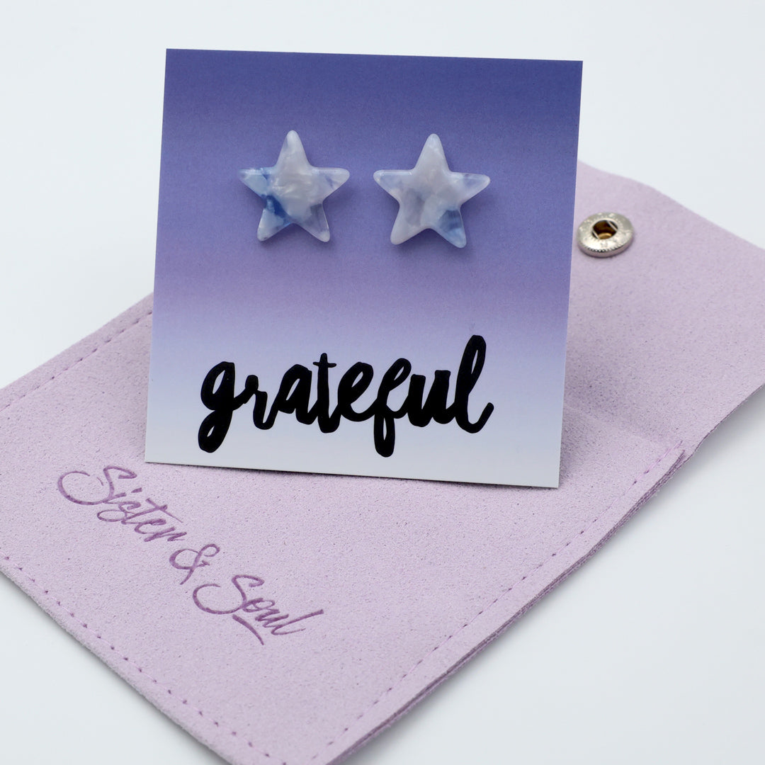 Two star-shaped stud earrings with a white and light blue marbled pattern, displayed on a purple gradient card that says 'grateful'. The card is resting on a lavender fabric pouch with 'Sister & Soul'.