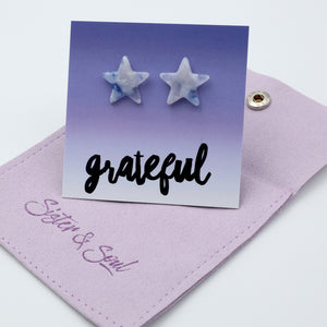 Two star-shaped stud earrings with a white and light blue marbled pattern, displayed on a purple gradient card that says 'grateful'. The card is resting on a lavender fabric pouch with 'Sister & Soul'.