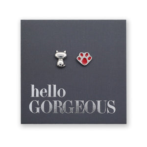 A pair of small stud earrings displayed on a dark gray backing card. One earring is a silver cat-shaped charm, and the other is a red paw print earring.