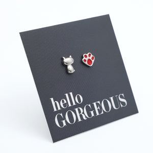 A pair of pet-themed stud earrings on a dark gray display card. One earring is a small silver cat silhouette, and the other is a silver paw print with a red pad.