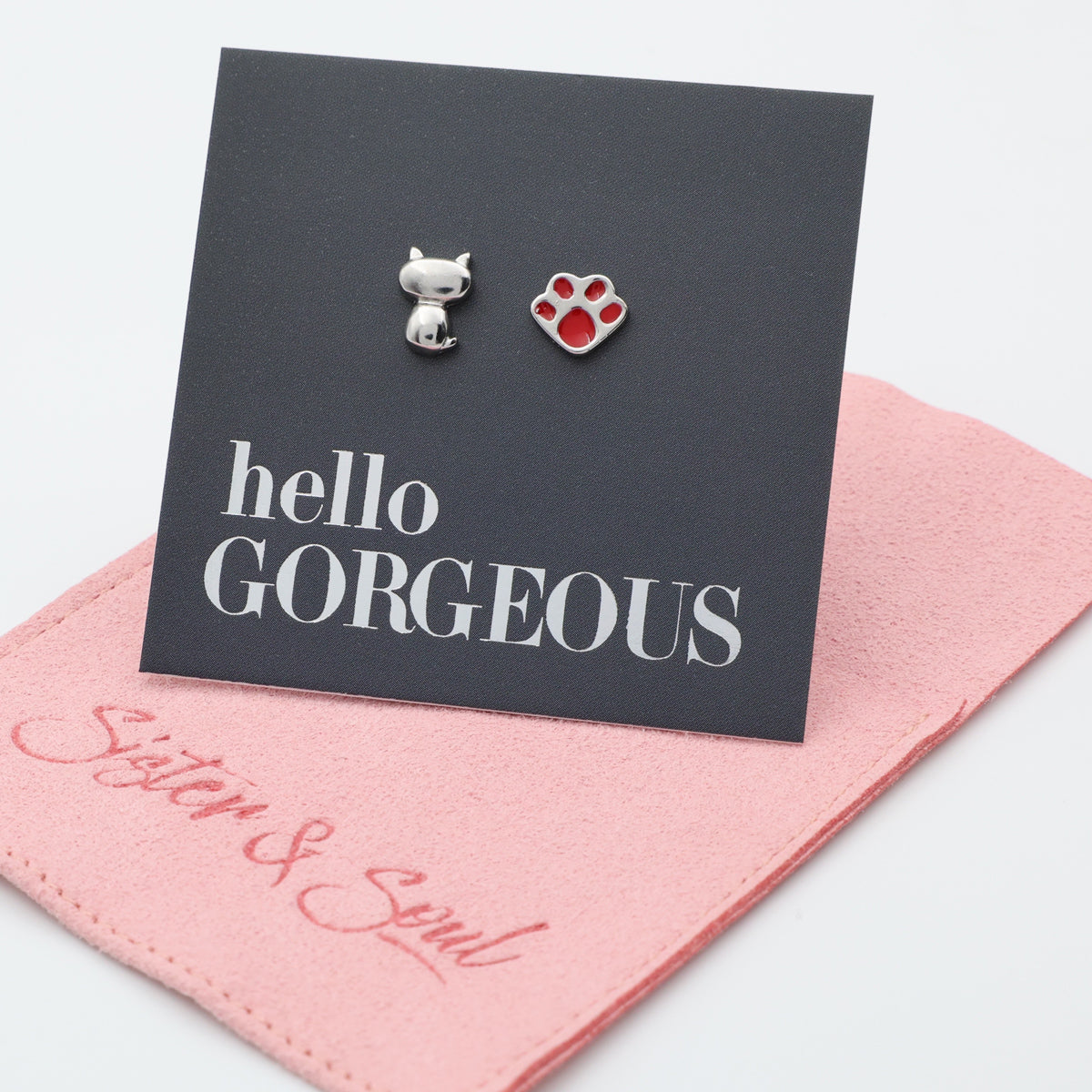 A silver cat-shaped stud and a silver paw-print stud with red accents stud earrings are displayed on a black card that reads 'hello GORGEOUS,' resting on a pink fabric pouch with 'Sister & Soul' wording. 