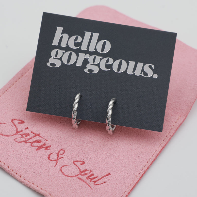 Silver twisty hoops on a grey card that has silver writing saying "Hello Gorgeous" on top of a pink pouch on a white background
