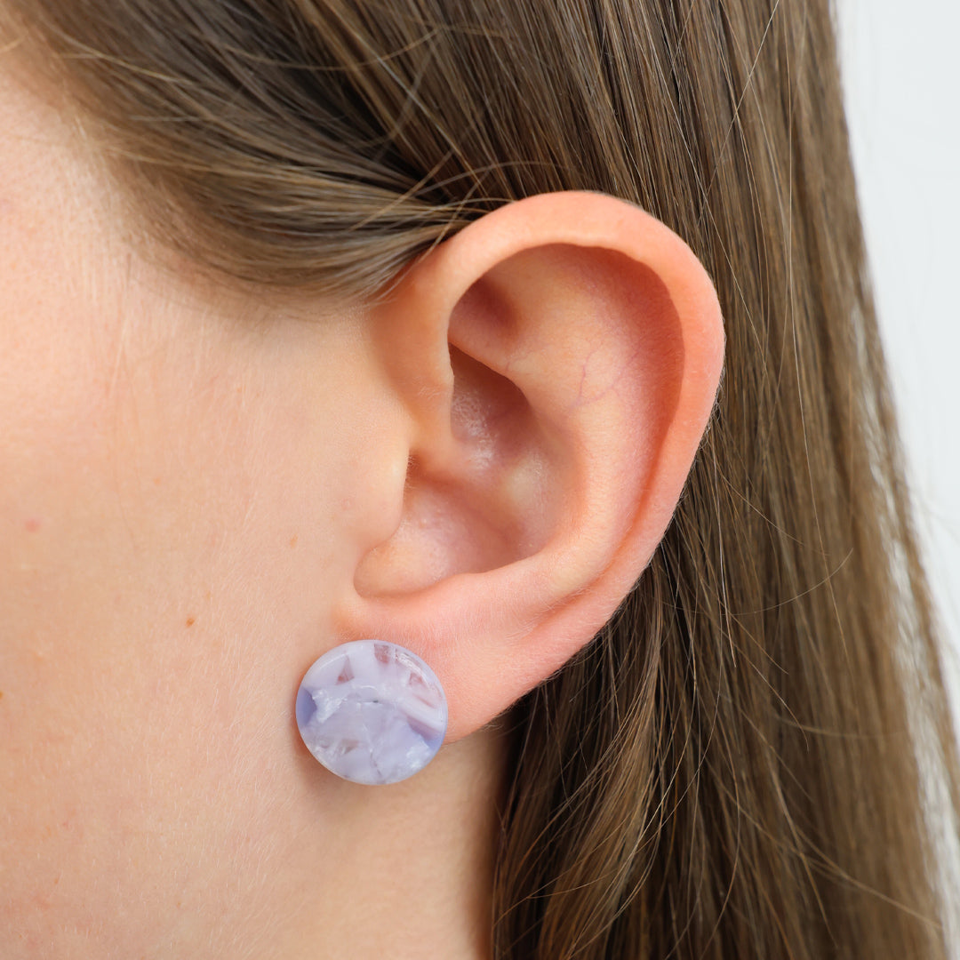 A close-up of a person's ear wearing a round stud earring with a marbled lavender and white pattern.