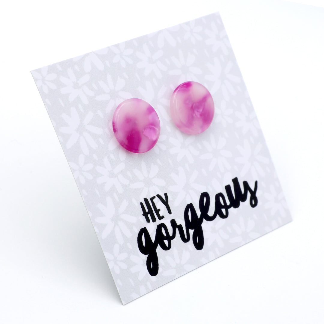 A pair of round stud earrings with a pink and purple marbled pattern, displayed on a white card with a floral pattern and the text 'HEY gorgeous' in black script.