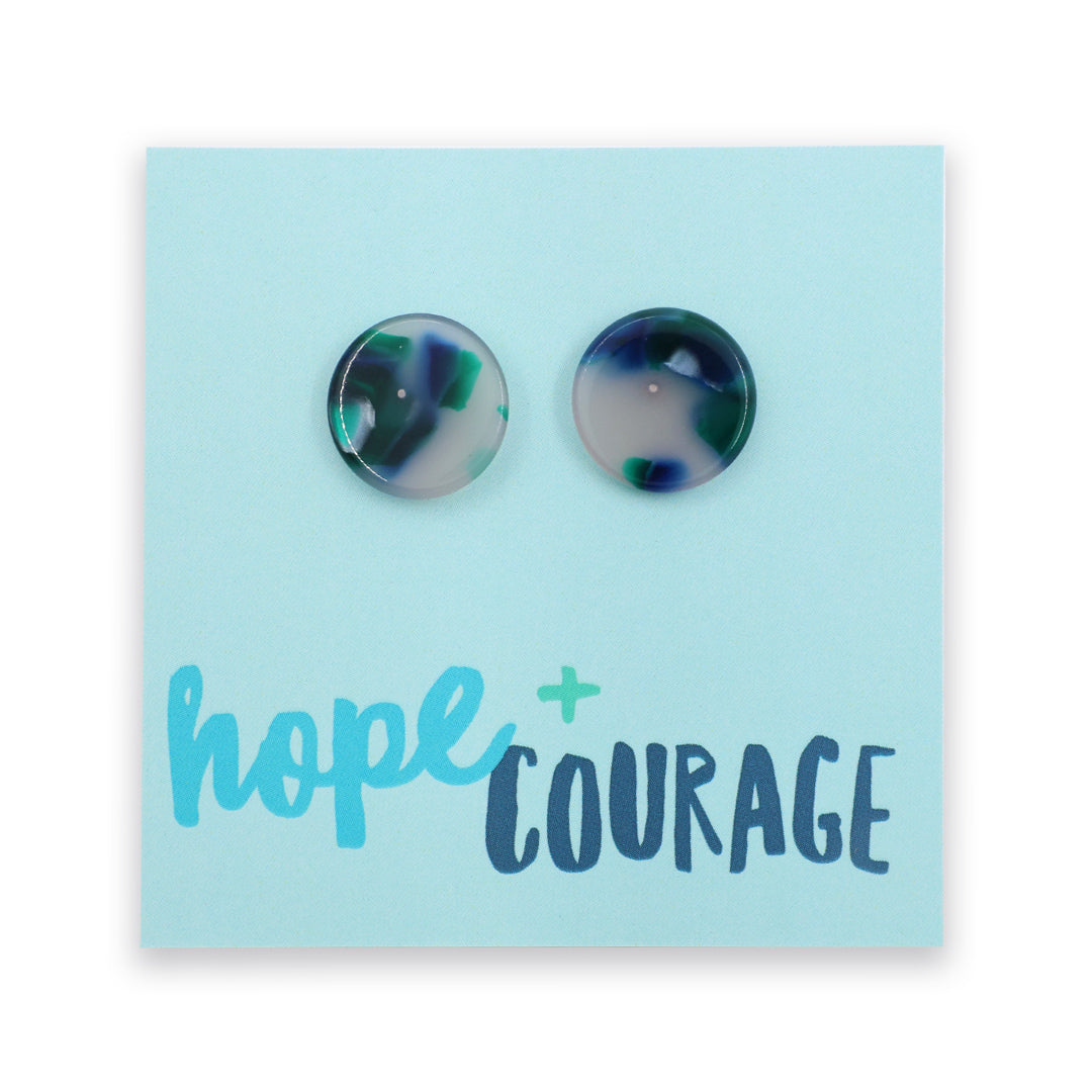 A pair of round marbled stud earrings with green, blue, and white swirls, displayed on a light blue backing card that reads 'hope + COURAGE'.