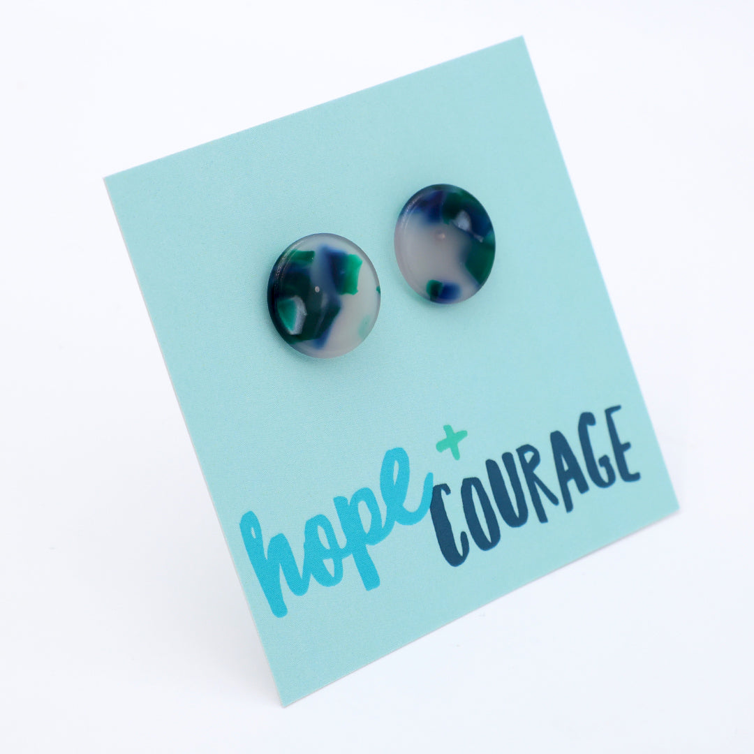 A pair of round stud earrings with a marbled dark blue, green, and gray pattern, mounted on a light blue backing card printed with the words 'hope + COURAGE'.