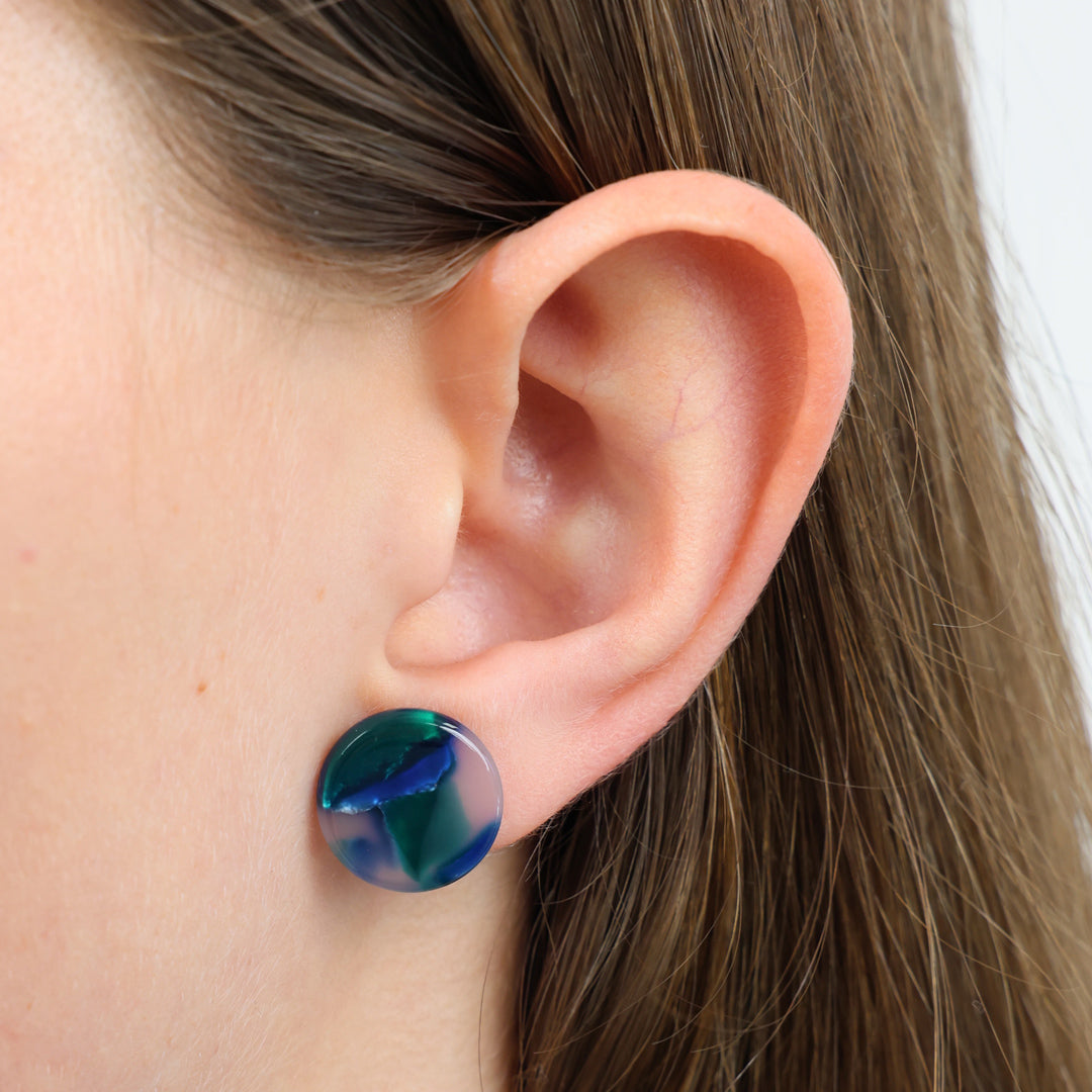 A close-up of a person's ear wearing a round stud earring with a marbled blue, green pattern.