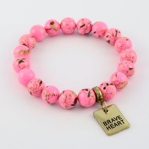 Pink beaded bracelet with a gold 'Brave Heart' charm on a light pink background