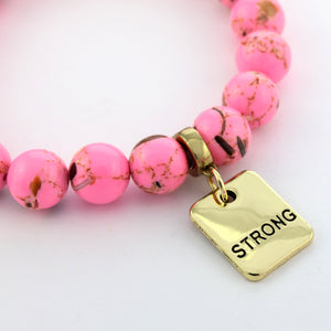 Pink beaded bracelet with a gold tag that says 'STRONG' on a white background
