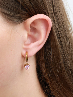 A close-up of a person's ear wearing a small gold hoop earring with a dangling heart-shaped pink gemstone.