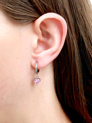 A close-up of a person's ear wearing a silver huggie hoop earring with a dangling pink heart charm.