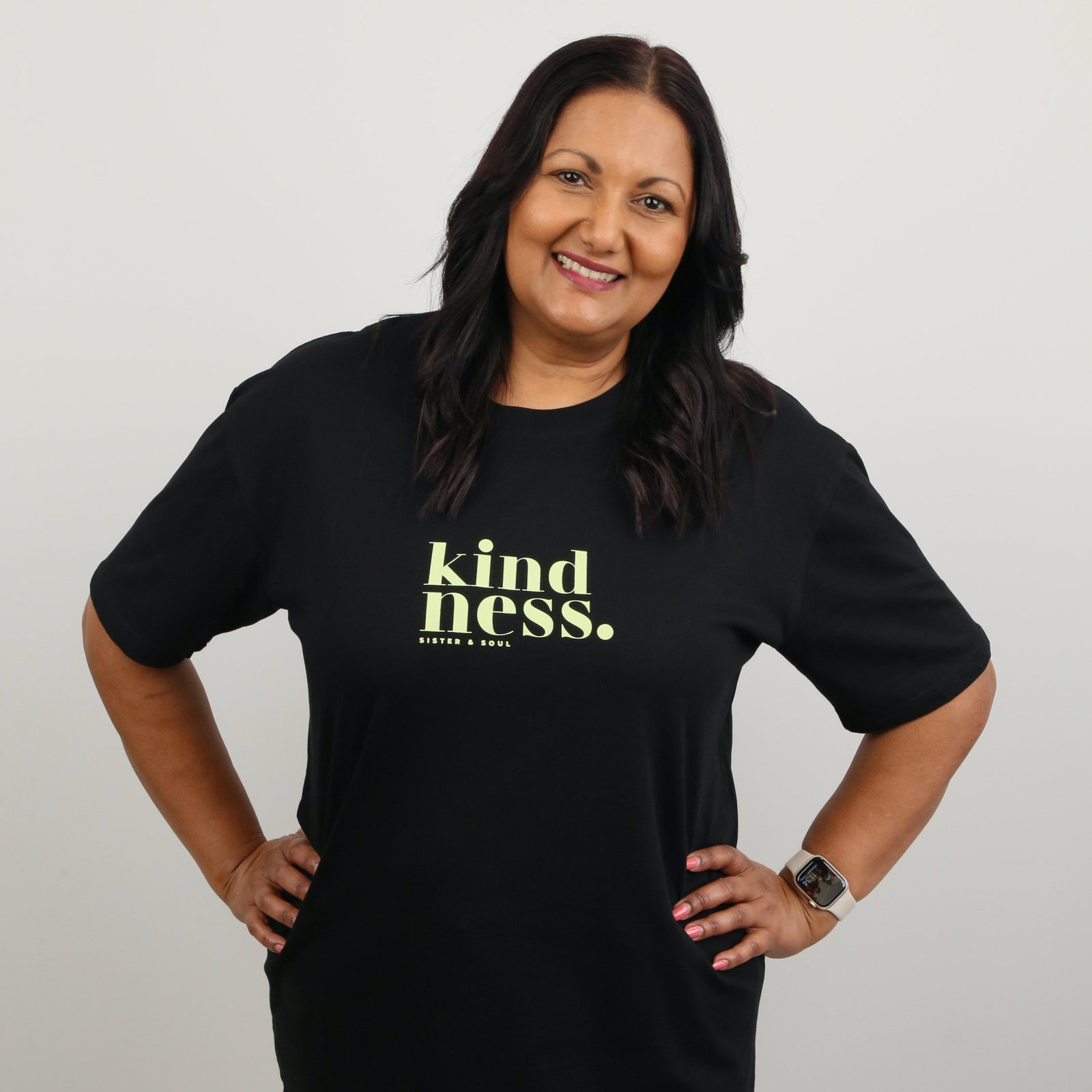 A smiling woman with dark hair stands with hands on hips wearing a black short-sleeve t-shirt with 'kindness.' printed in yellow-green text on the chest, paired with light blue jeans, against a plain light gray background.