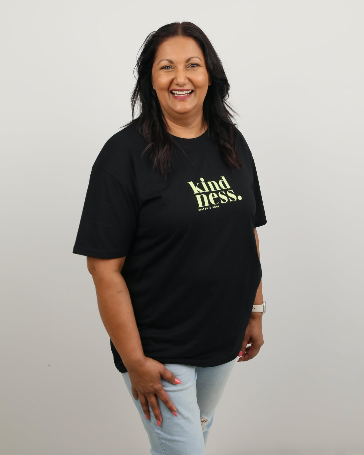 A smiling woman with dark hair stands against a plain light gray wall wearing a black short-sleeve t-shirt with 'kindness.' printed in yellow-green text on the chest, paired with light blue jeans.