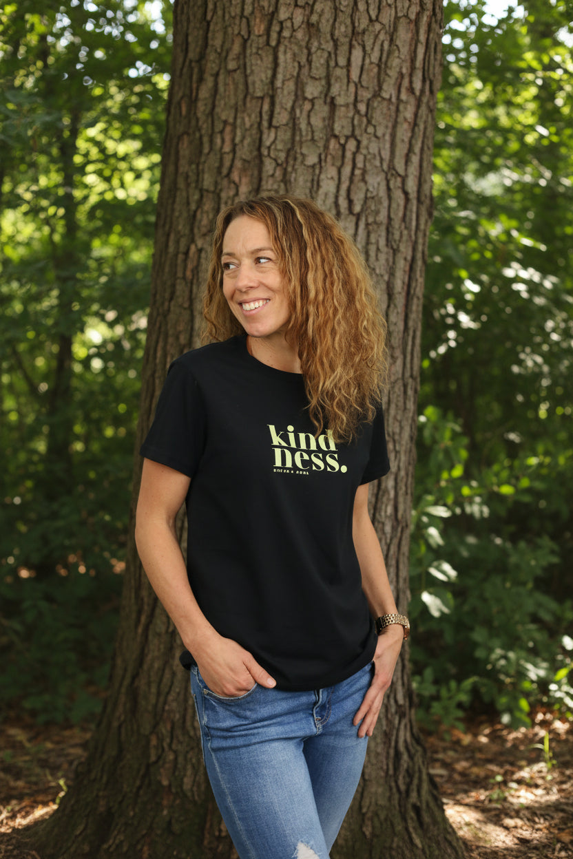 A woman with curly light brown hair wearing a black t-shirt with a yellow-green graphic that reads 'kindness. 