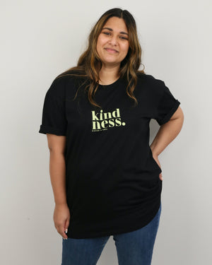 A woman wearing a black short-sleeve t-shirt with the word 'kindness.' printed in a pale yellow-green serif font on the chest, paired with blue jeans, standing against a plain light gray wall.