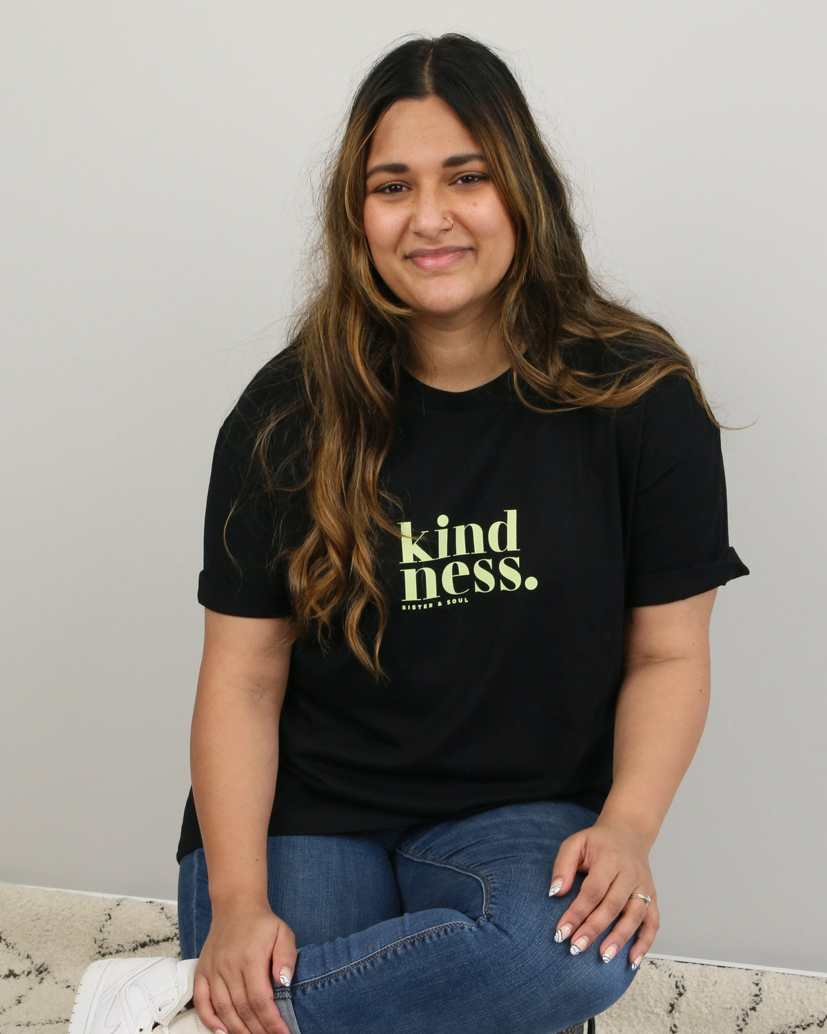 A woman sitting on a dark round ottoman wearing a black graphic t-shirt with the text 'kindness.' in yellow-green, blue jeans, and white sneakers, set against a plain light gray wall and a patterned rug.