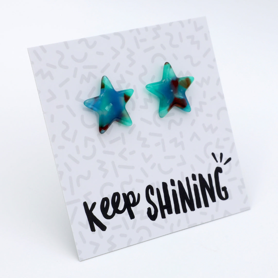 Two star-shaped stud earrings with a marbled blue, teal, and dark brown pattern, displayed on a white card that reads 'Keep SHINING'.