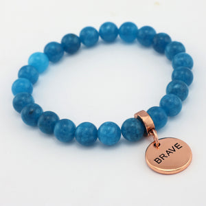 Blue beaded bracelet with a rose gold charm labeled 'BRAVE' on a white background