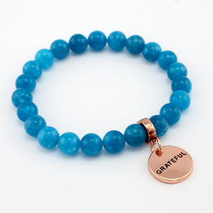Blue beaded bracelet with a rose gold 'GRATEFUL' charm on a white background